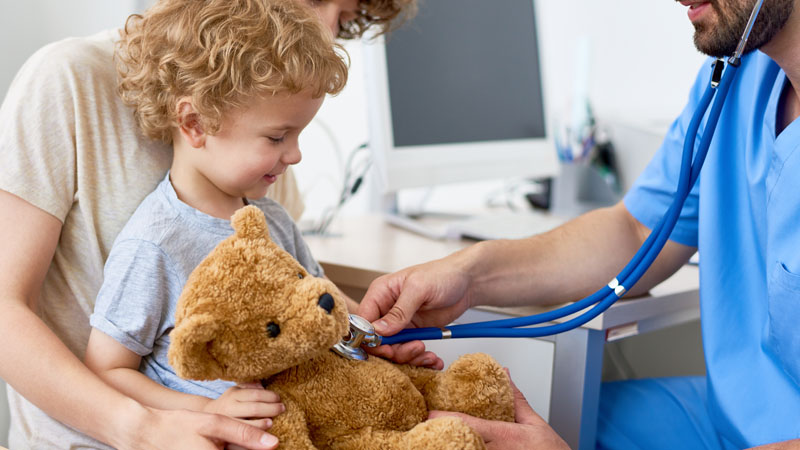 Portrait of adorable curly child  sitting on mothers lap in doctors office holding teddy bear toy, with pediatrician listening to heartbeat using stethoscope